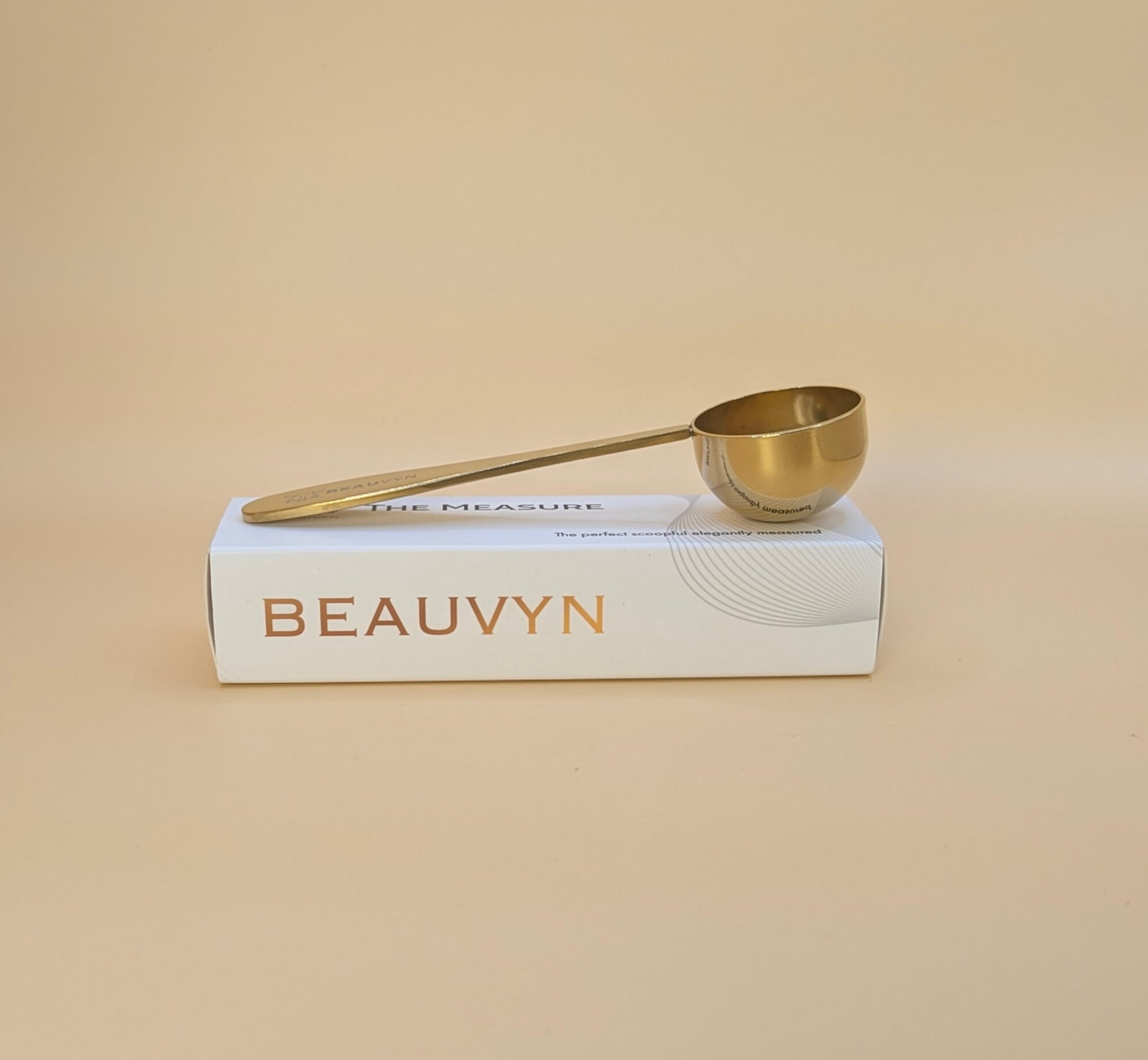 Gold measuring spoon on a box with 'BEAUVYN' branding against a beige background